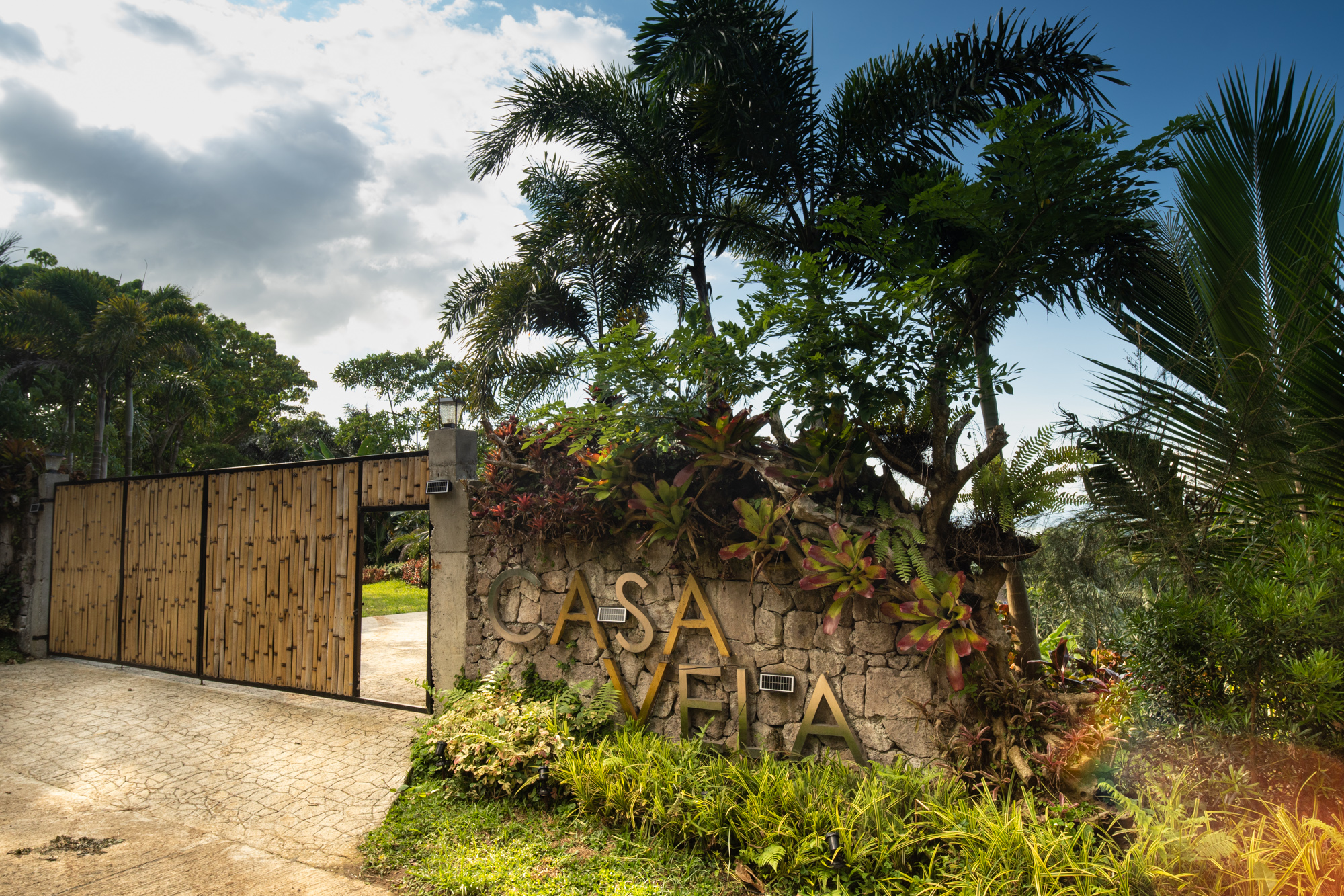 Casa Vela entrance with stone wall sign, tropical gate, and driveway under a partly cloudy sky