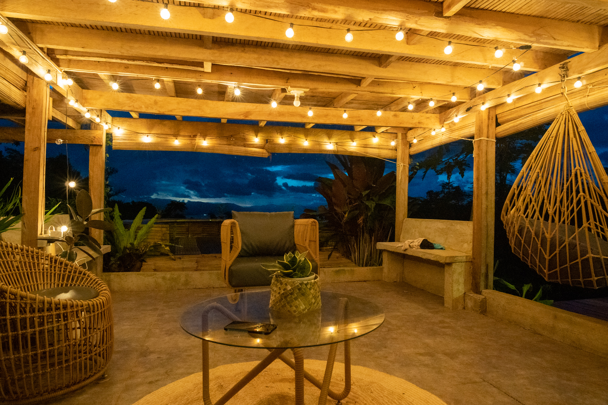 Cozy boho-style vacation rental patio with string lights, wicker seating, hanging chair, glass coffee table, tropical plants, and scenic sunset mountain view