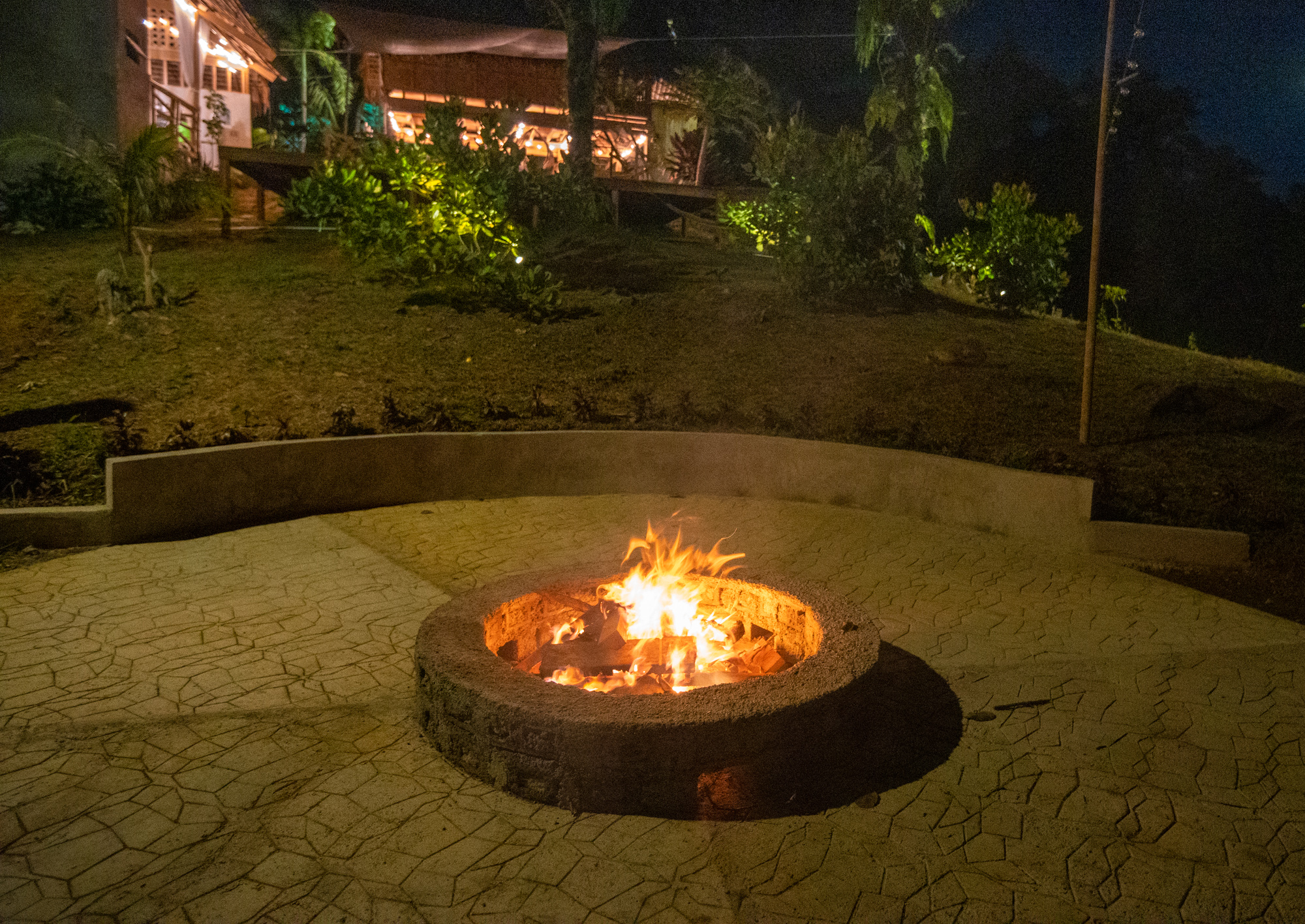 Nighttime tropical vacation rental patio with a round stone fire pit, warm garden lighting, lush landscaping, and a glowing lodge-style house in the background