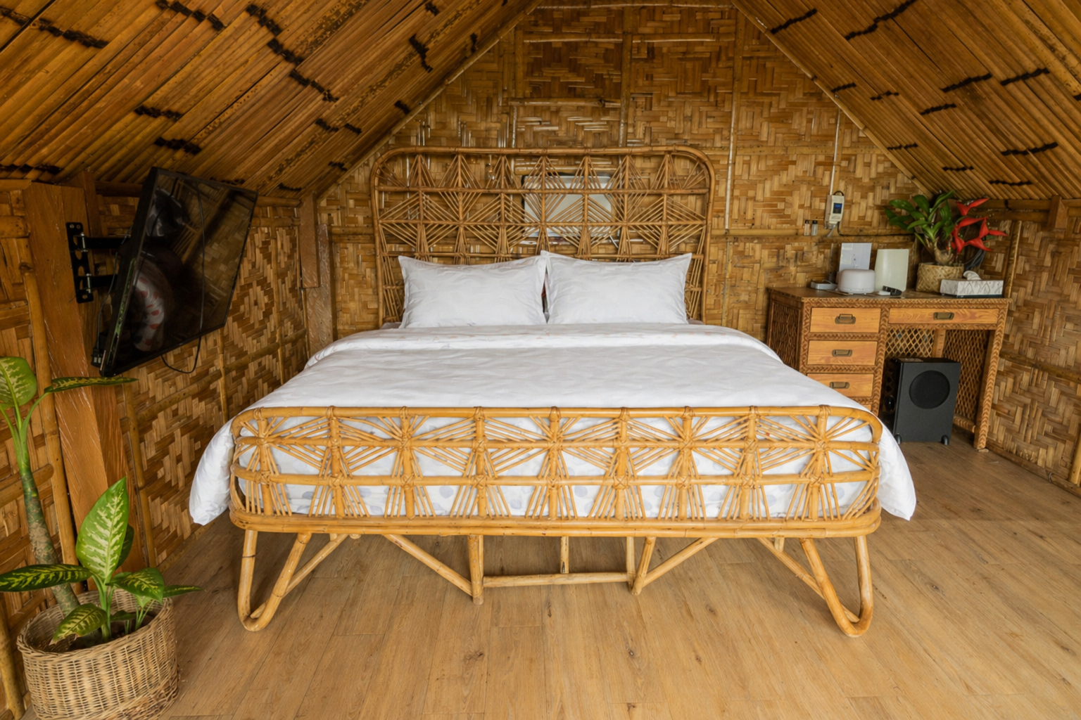 Rustic bamboo bedroom in a tropical vacation rental with a rattan bed, white bedding, woven walls, thatched ceiling, wall-mounted TV, and wooden desk
