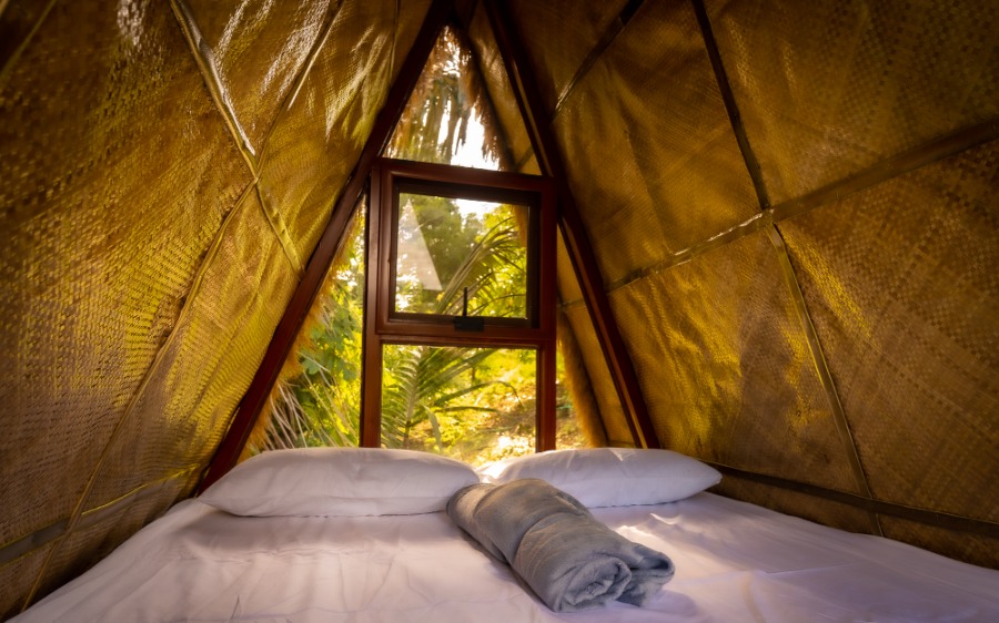 Interior of an A-frame glamping cabin with a bed, white linens, and tropical greenery visible through the window, ideal for a secluded vacation rental getaway