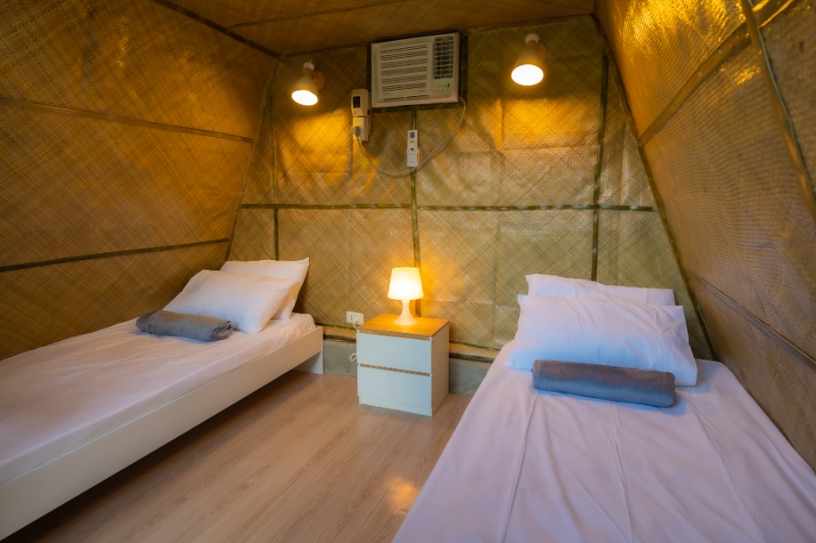 Cozy glamping-style bedroom with two single beds, white linens, bedside lamp, wall-mounted air conditioner, and warm wood flooring inside a vacation rental cabin