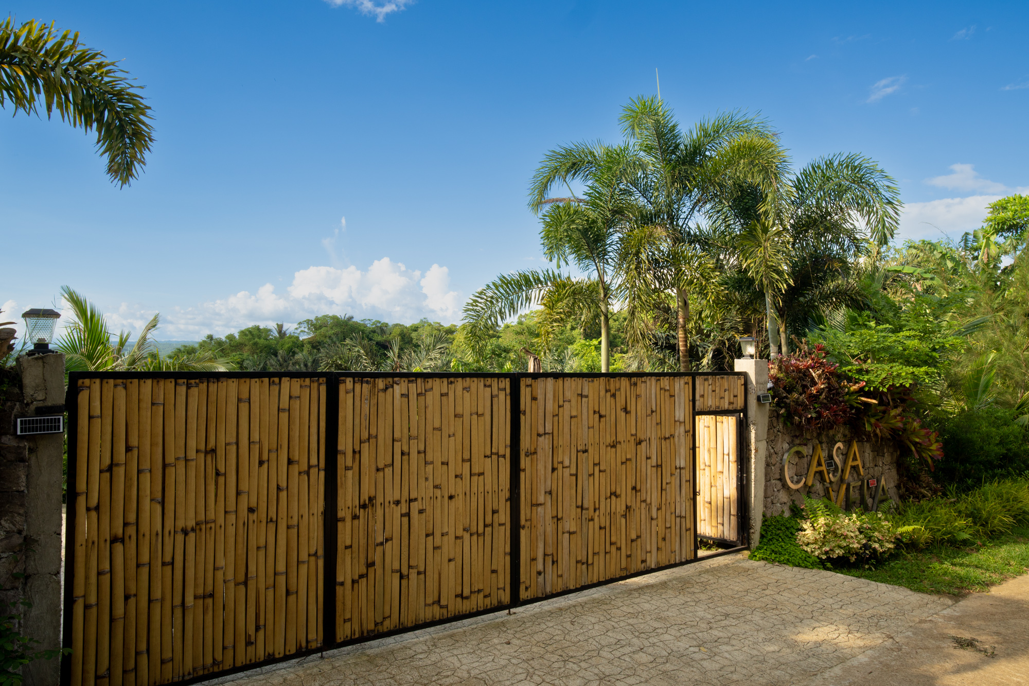 Tropical vacation rental entrance with bamboo gate, lush palm landscaping, and clear blue sky