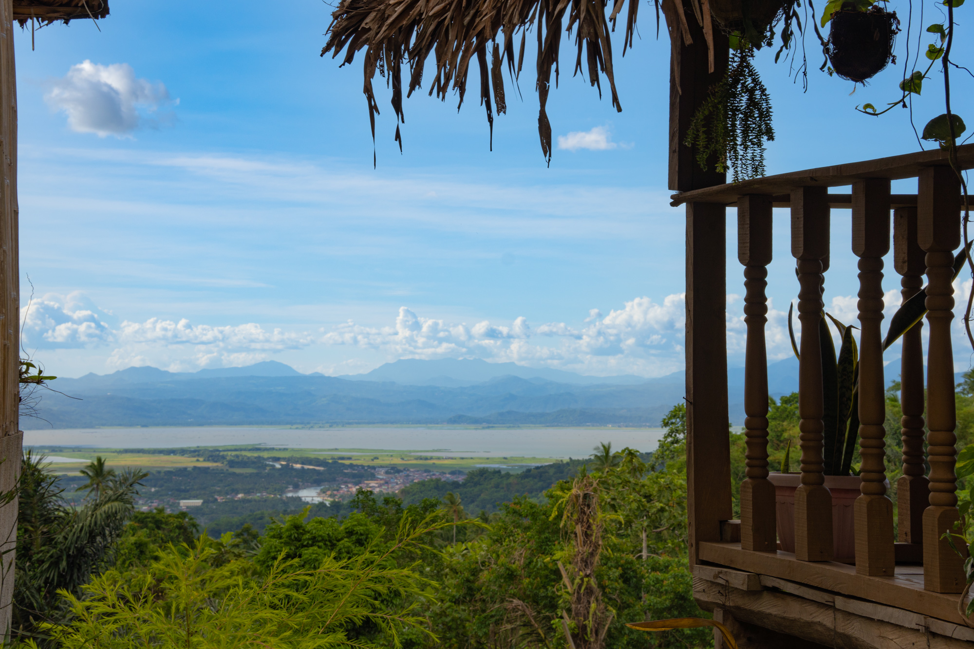 Rustic tropical vacation rental balcony with wooden railing overlooking lush green hills, a coastal town, calm water, and distant mountains under a blue sky with clouds