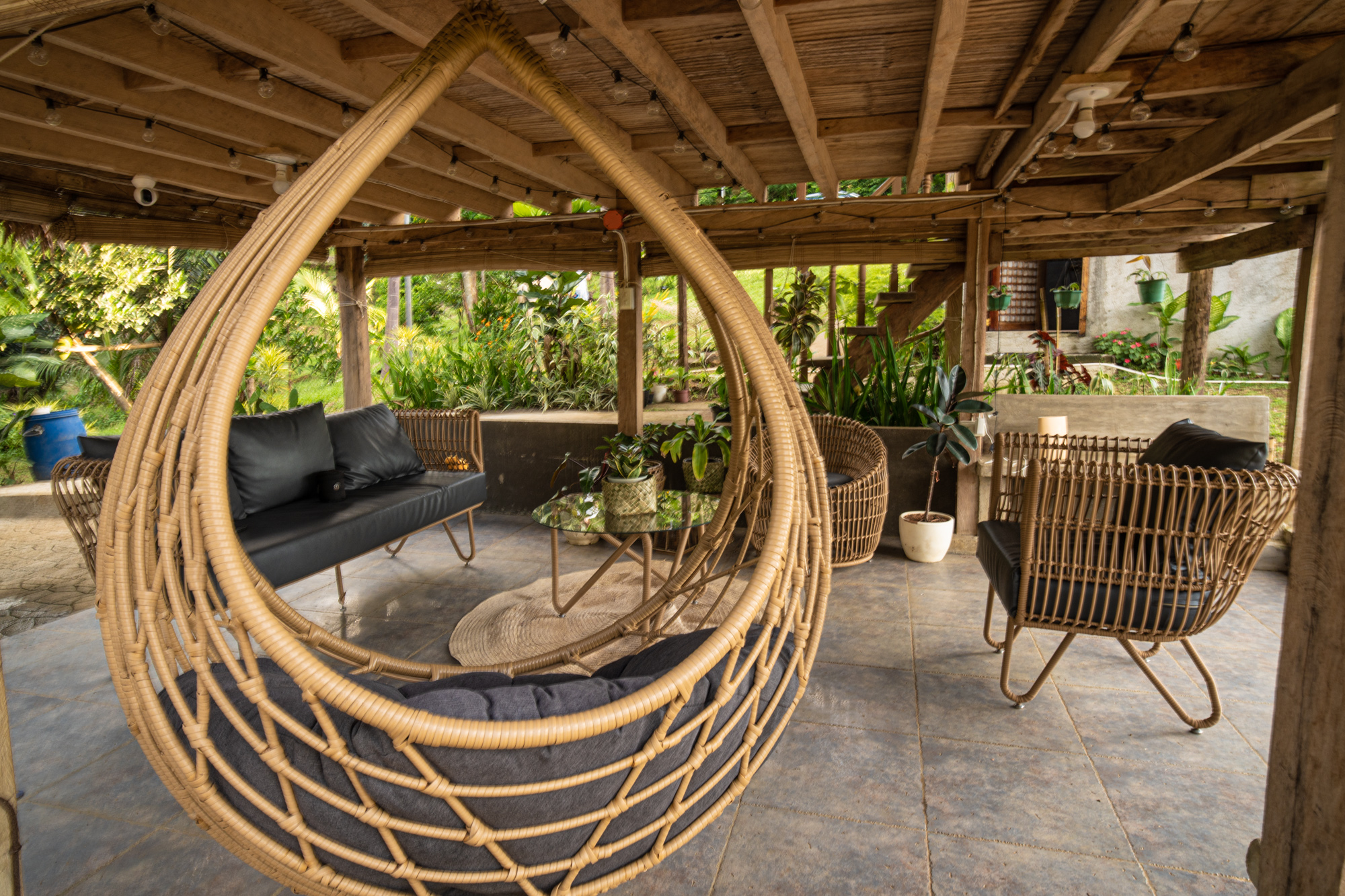 Covered tropical outdoor lounge with wicker seating, hanging chair, glass coffee table, and panoramic ocean and mountain views from a vacation rental terrace