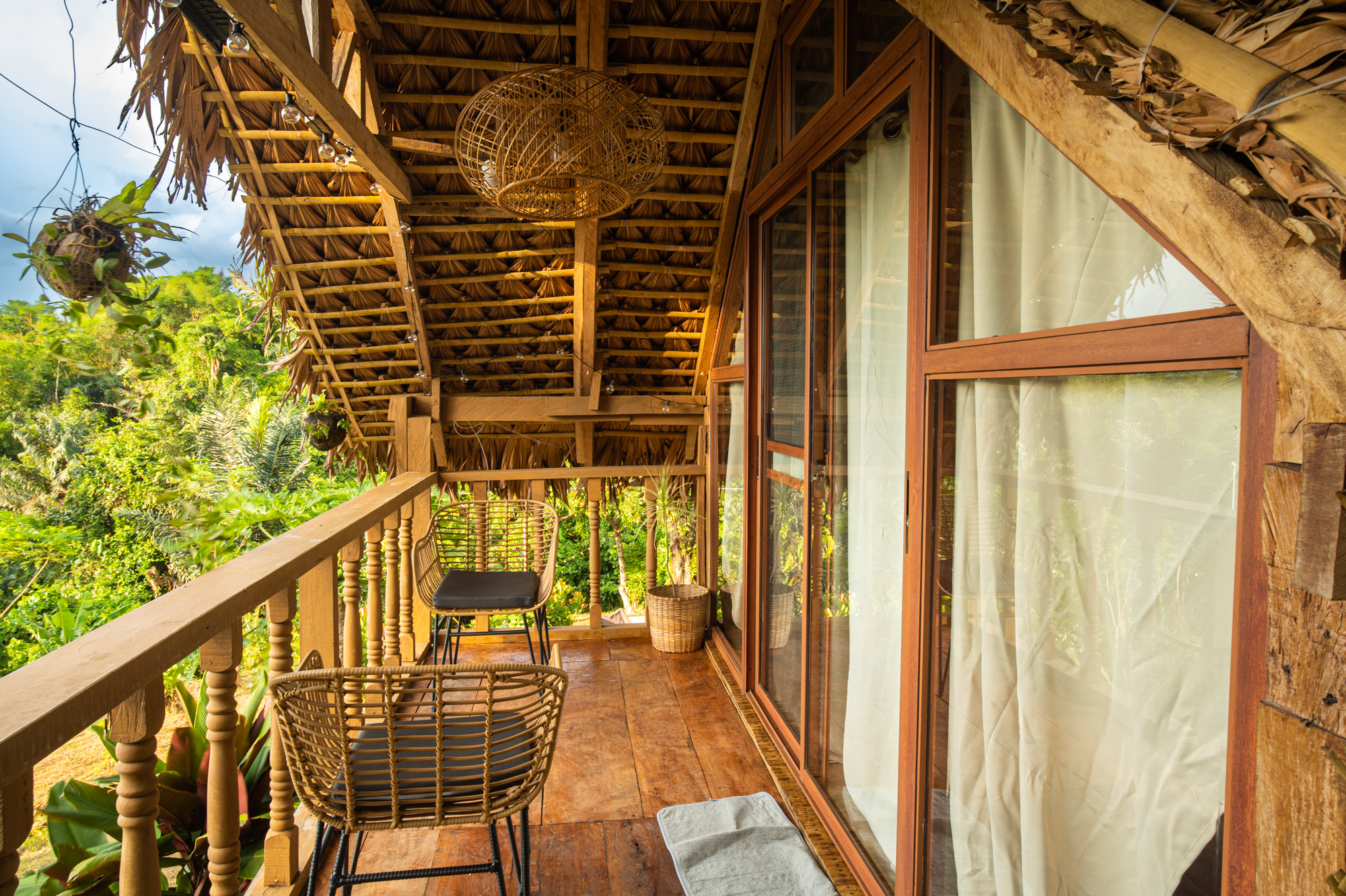 Rustic tropical vacation rental balcony with wicker chairs, wooden deck, thatched roof, sliding glass doors, and lush jungle views