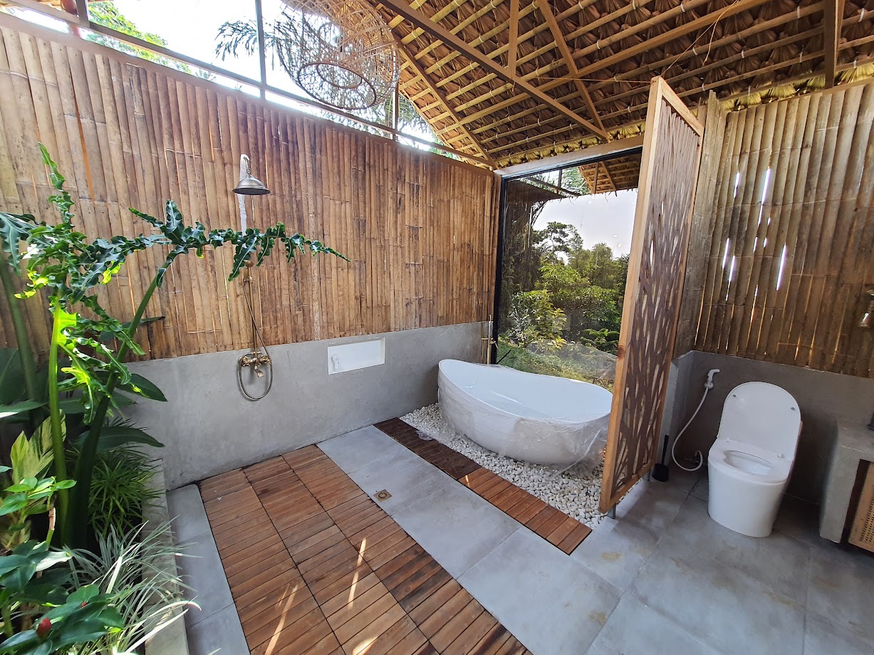 Tropical open-air bathroom in a vacation rental with a freestanding soaking tub, bamboo walls and thatched roof, wooden floor panels, lush greenery, and a modern toilet in a private jungle-inspired spa setting
