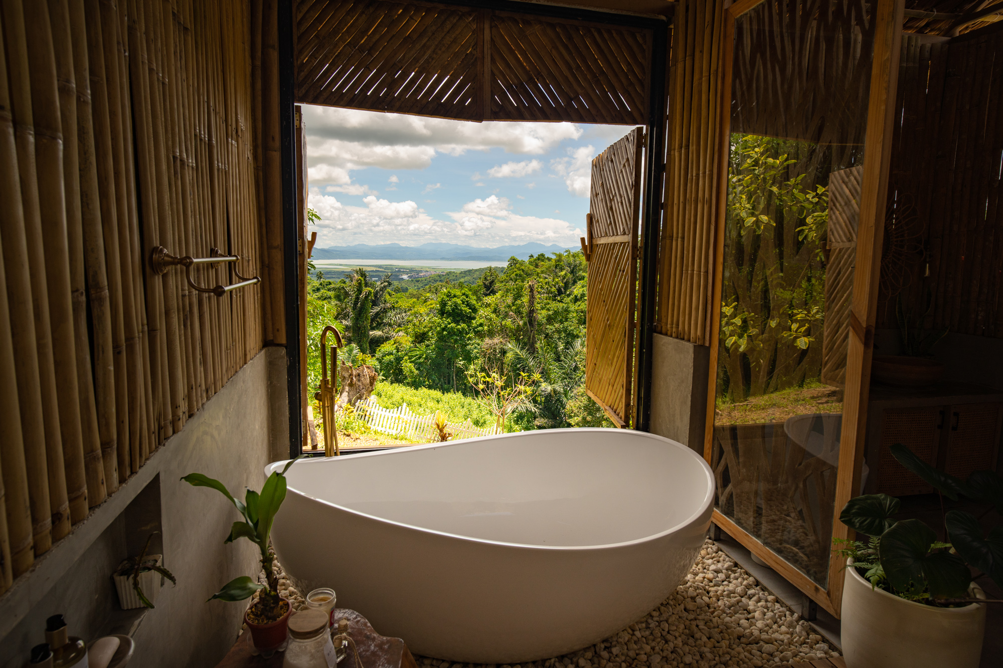 Open-air tropical bathroom with a freestanding white soaking tub, bamboo walls, pebble floor, and panoramic jungle, mountain, and ocean view in a luxury vacation rental